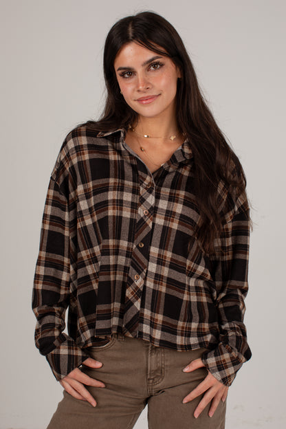 A woman modeling a long-sleeve button-up plaid shirt in black, brown, and beige, paired with brown pants, against a plain light gray studio background.