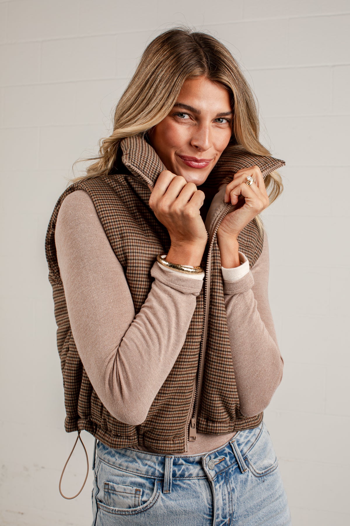 A woman wearing a brown and beige houndstooth zip-up vest over a beige long-sleeve top and blue jeans, posing against a white paneled wall.