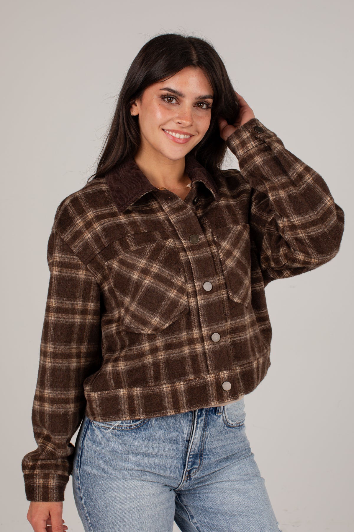 A woman wearing a brown and beige plaid cropped flannel shirt jacket with a solid brown corduroy collar, paired with light blue jeans, against a plain light gray background.