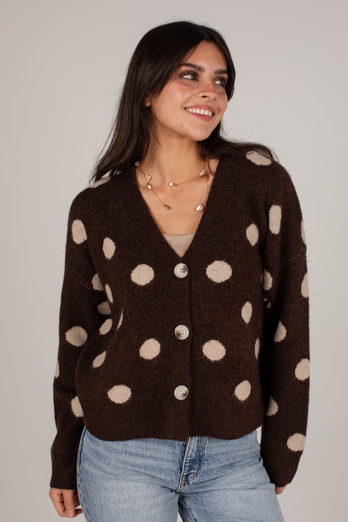 A woman wearing a dark brown knit cardigan with beige polka dots and silver buttons, paired with light blue jeans, against a plain light gray background.