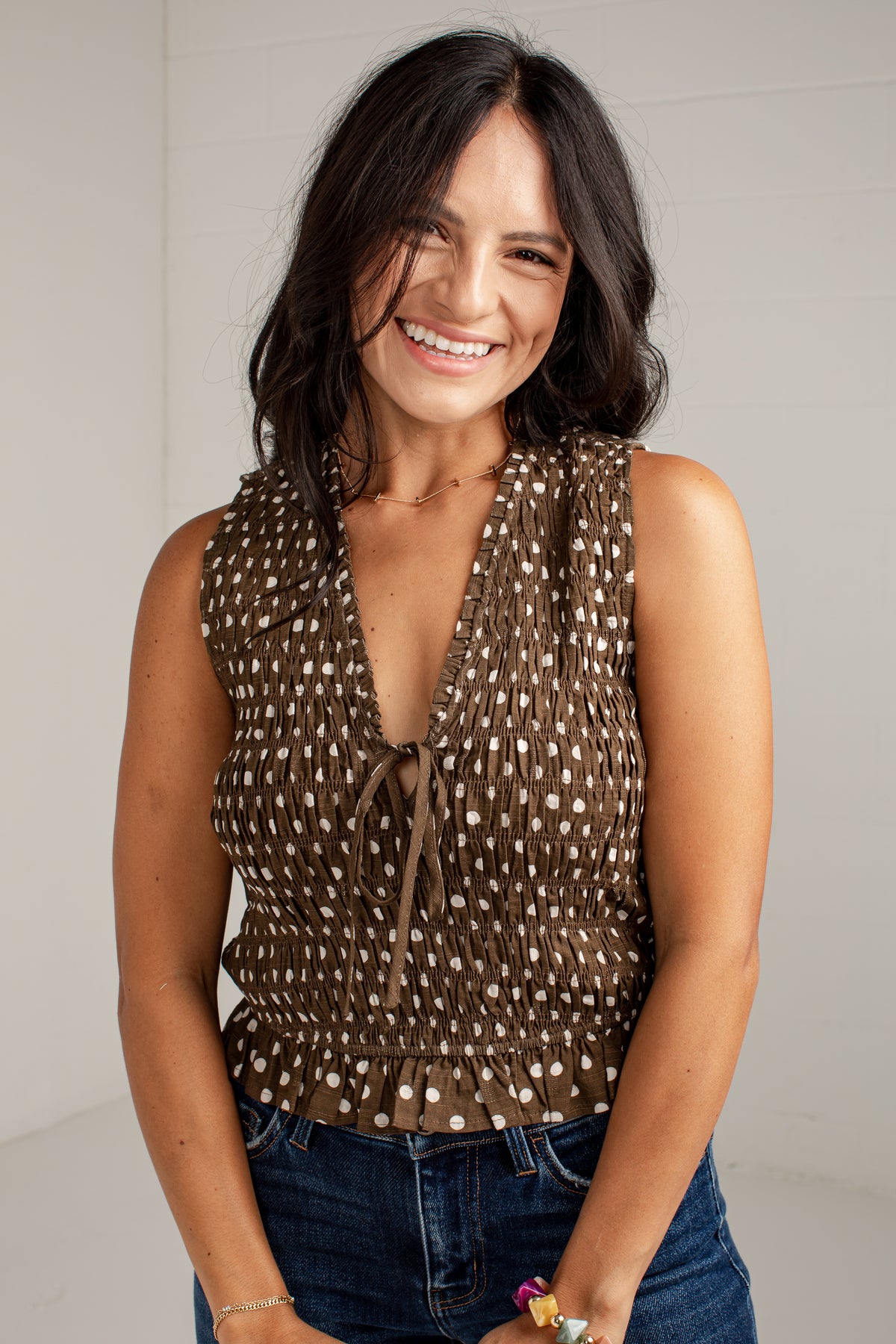 A smiling woman with dark hair wearing a brown sleeveless top with a white polka-dot pattern and tie neckline, paired with blue jeans, against a light gray background.