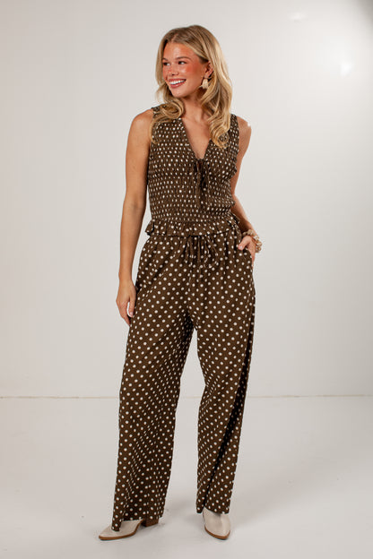 A woman modeling a sleeveless brown jumpsuit with a white polka dot pattern, featuring a tie-neck detail and wide legs, set against a plain white studio background.