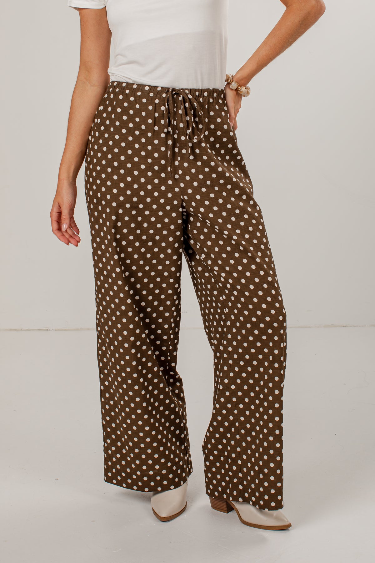 A person wearing brown wide-leg pants with a white polka dot pattern, styled with a white top and beige heeled sandals against a plain white studio background.