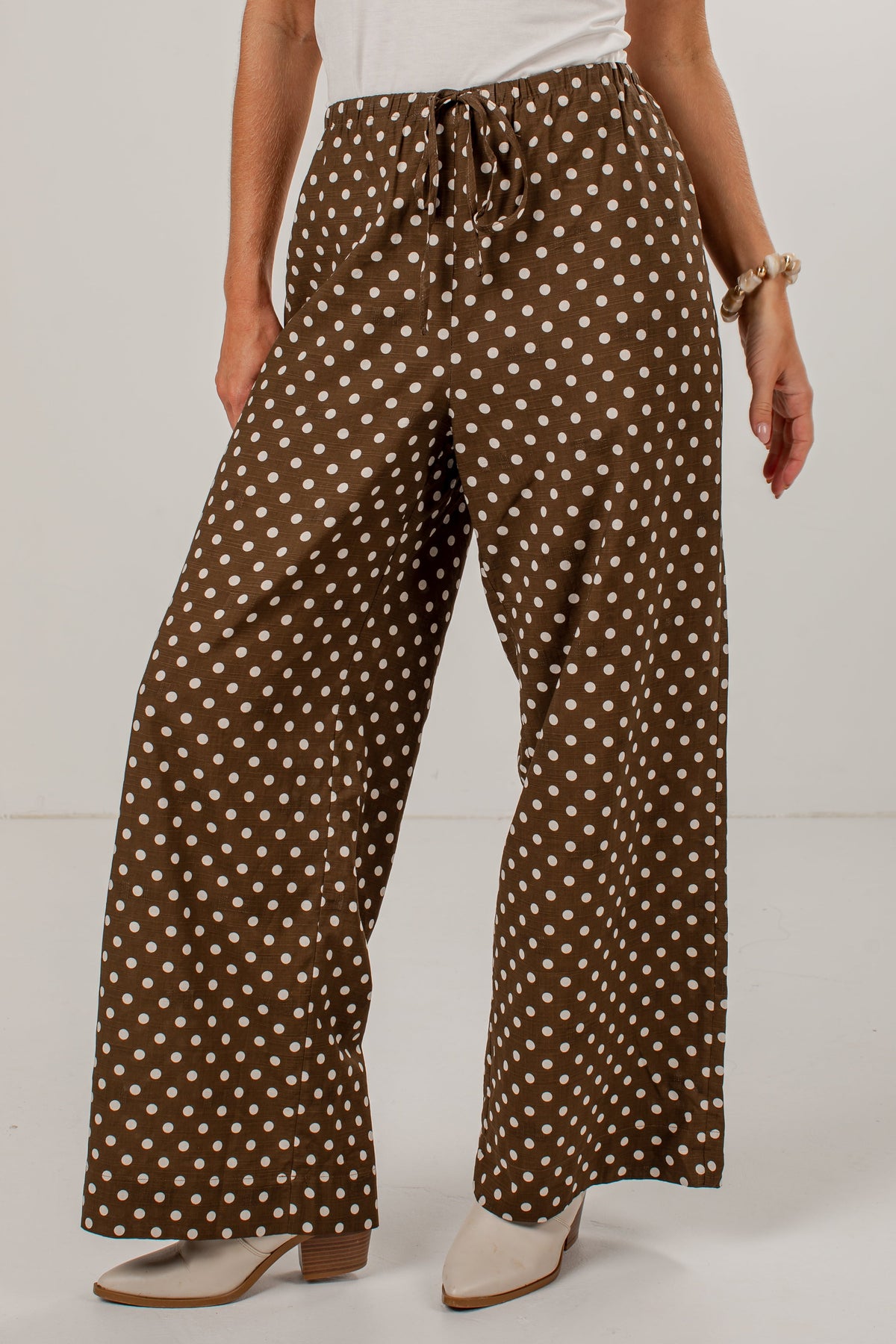 A person wearing brown wide-leg pants with a white polka dot pattern, styled with a white top and beige heeled shoes against a plain white studio background.