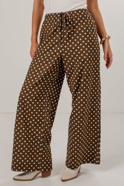A person wearing brown wide-leg pants with a white polka dot pattern, styled with a white top and beige heeled shoes against a plain white studio background.
