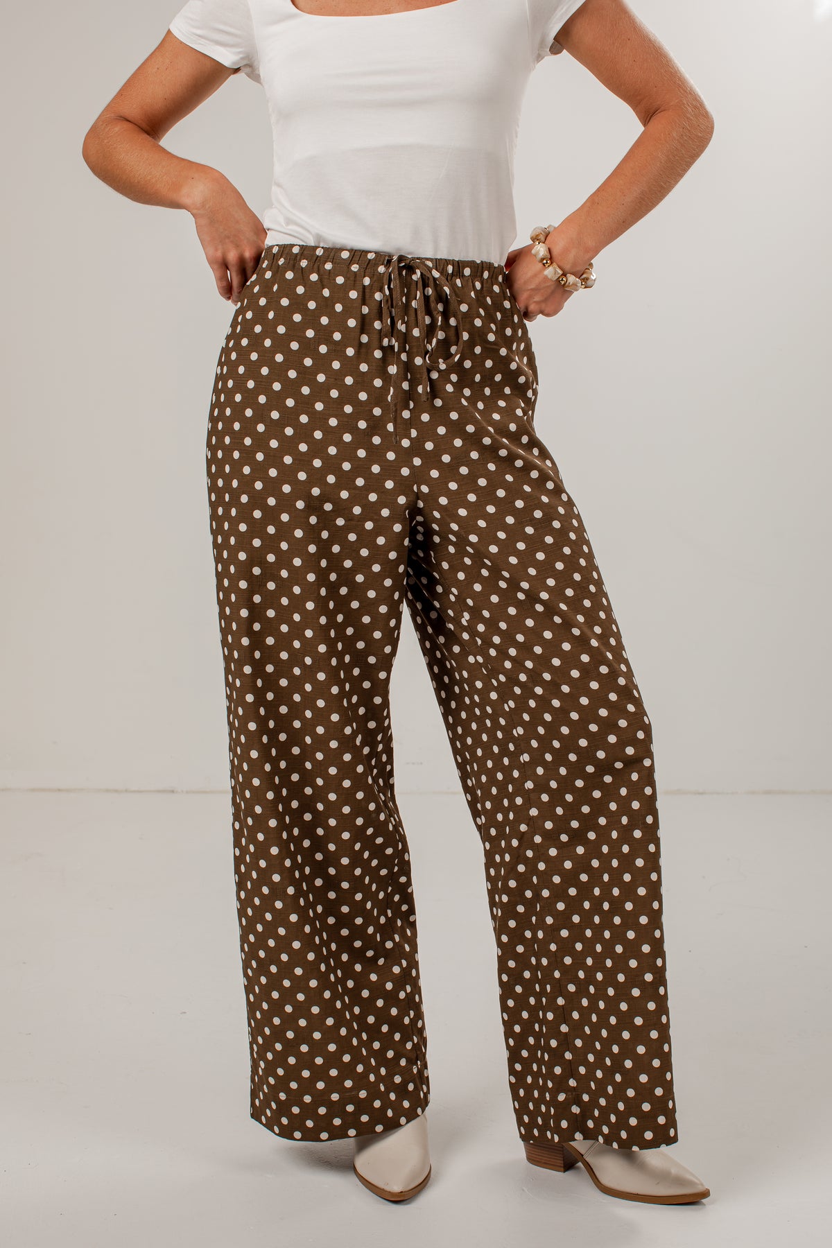 A model wearing brown wide-leg pants with a white polka dot pattern, styled with a white top and white pointed-toe heels against a plain light gray studio background.