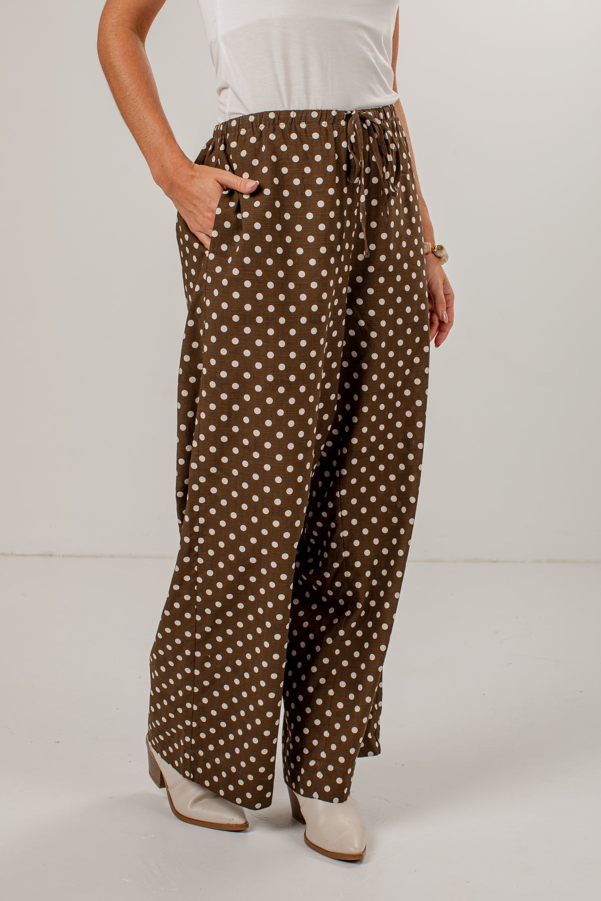 A person wearing loose brown wide-leg pants with a white polka dot pattern, paired with a white top and white heeled boots, shown against a plain light gray studio background.