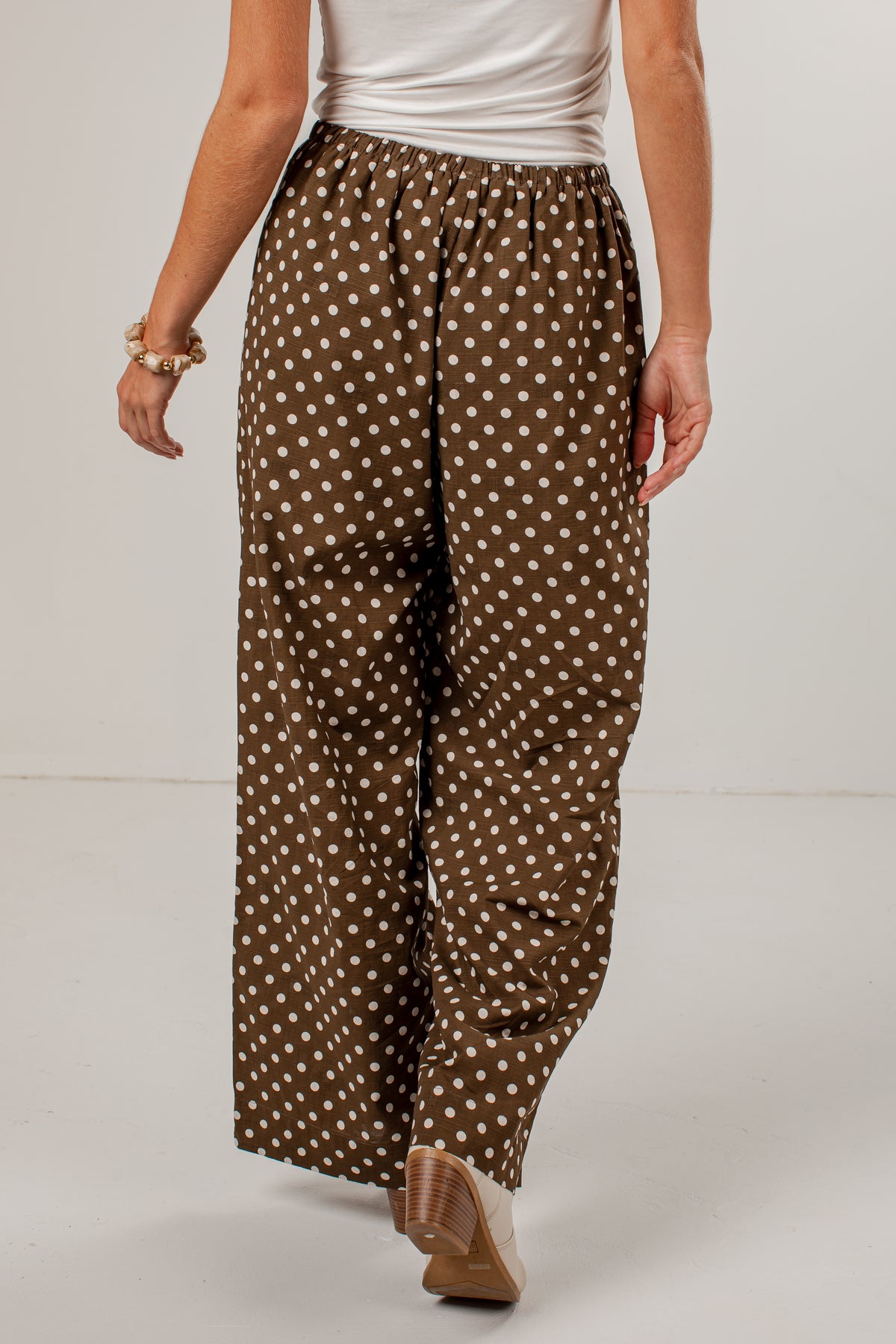 A person wearing loose brown wide-leg pants with a white polka dot pattern, paired with a white top and tan heels, shown from the waist down against a light gray studio background.