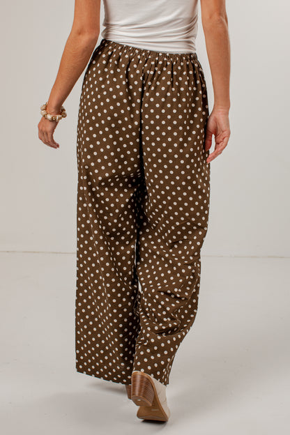 A person wearing loose brown wide-leg pants with a white polka dot pattern, paired with a white top and tan heels, shown from the waist down against a light gray studio background.