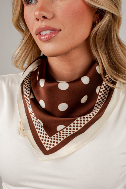 A close-up of a woman wearing a brown silk-like scarf with white polka dots and a geometric border, styled around her neck over a white top.