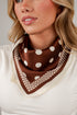 A close-up of a woman wearing a brown silk-like scarf with white polka dots and a geometric border, styled around her neck over a white top.