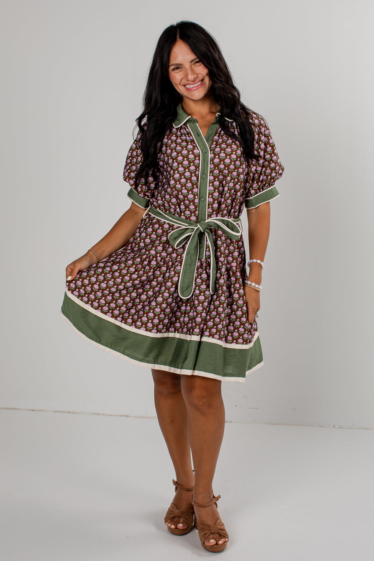 A woman modeling a short-sleeve button-front dress with an allover brown and white floral pattern, olive green trim, and a matching green tie belt, photographed against a plain light gray studio background.