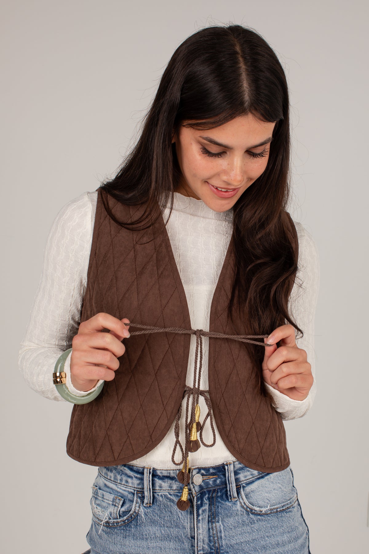 A woman wearing a brown diamond-quilted sleeveless vest over a white long-sleeve top and blue jeans, holding the front tie closure of the vest against a plain light gray background.