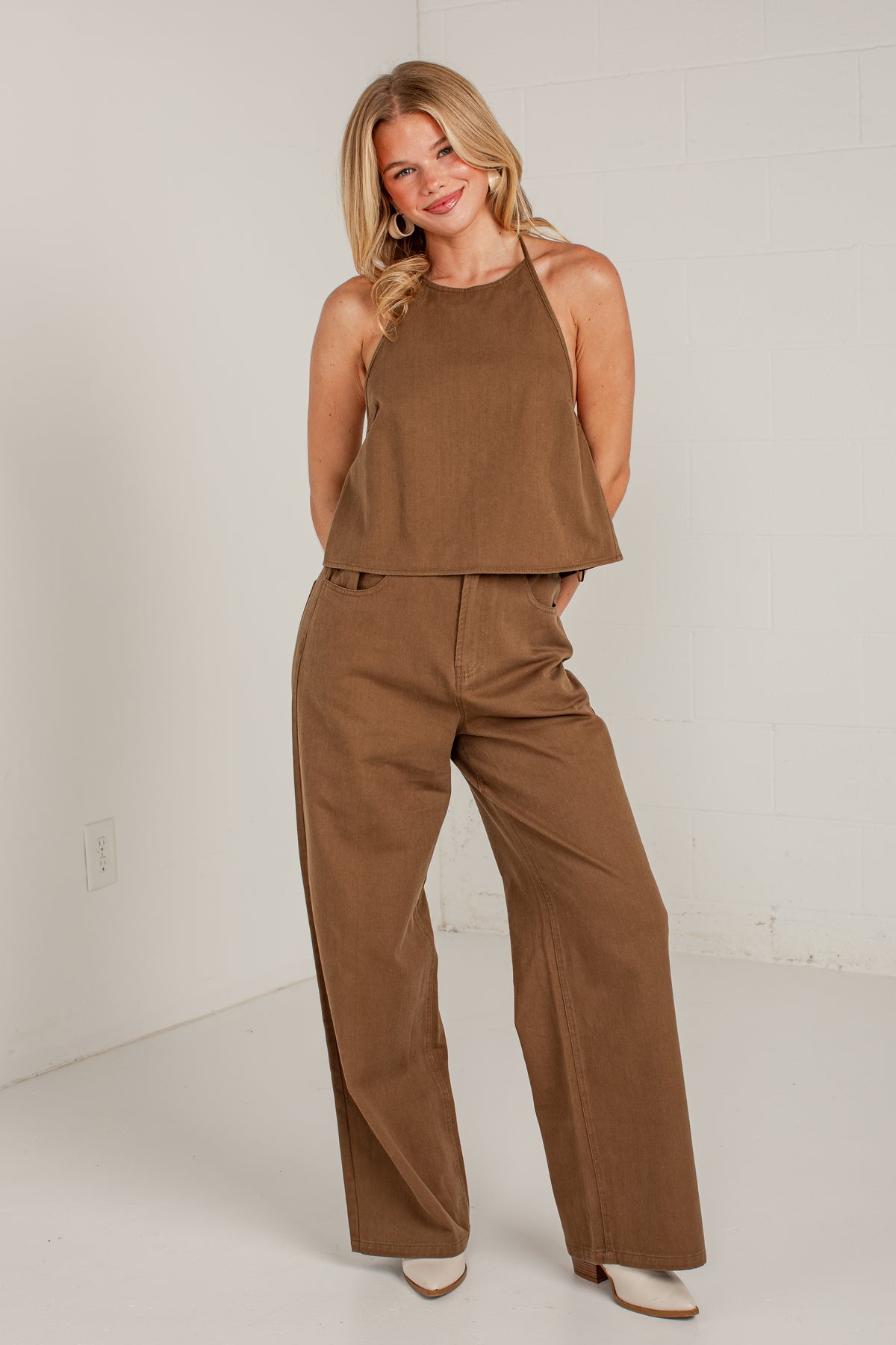 A woman modeling a two-piece brown corduroy set consisting of a sleeveless halter crop top and matching wide-leg pants, standing against a white brick wall.