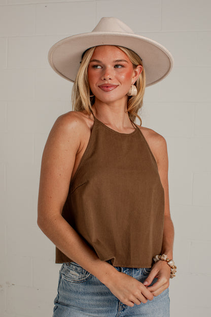 A woman wearing a brown halter crop top, blue jeans, and a wide-brimmed beige hat, standing against a light gray brick wall.