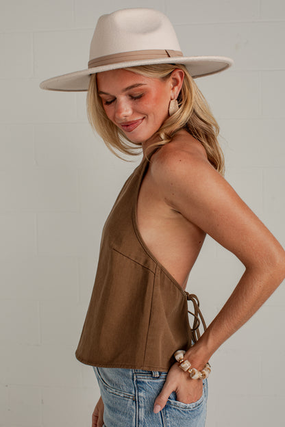 A woman wearing a brown halter-style sleeveless top with side tie details, blue jeans, and a wide-brimmed cream hat, photographed against a light gray wall.