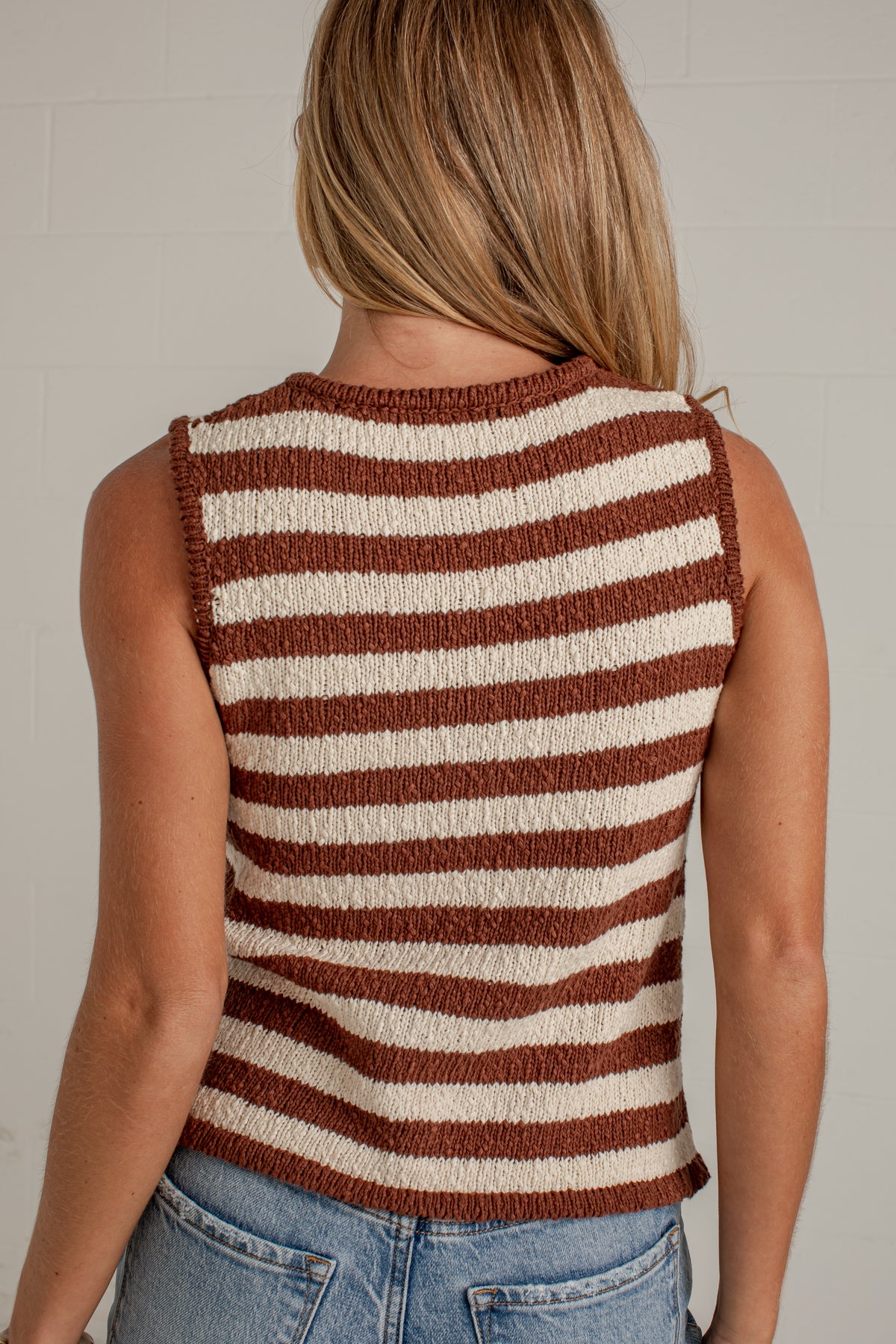 A woman viewed from the back wearing a sleeveless knit top with horizontal brown and cream stripes, paired with blue jeans against a light gray wall.
