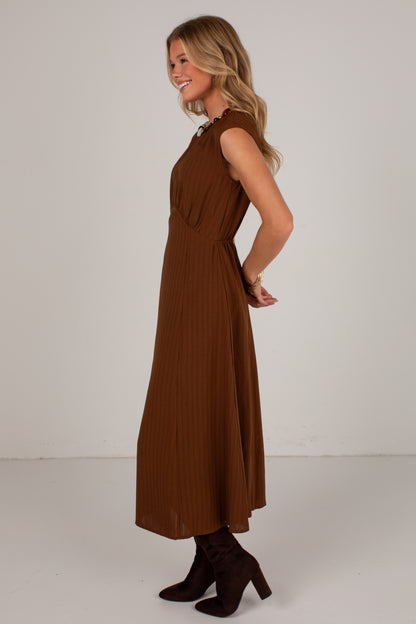 A woman modeling a sleeveless brown pleated midi dress with a gathered waist, paired with dark brown heeled boots, against a plain light gray studio background.