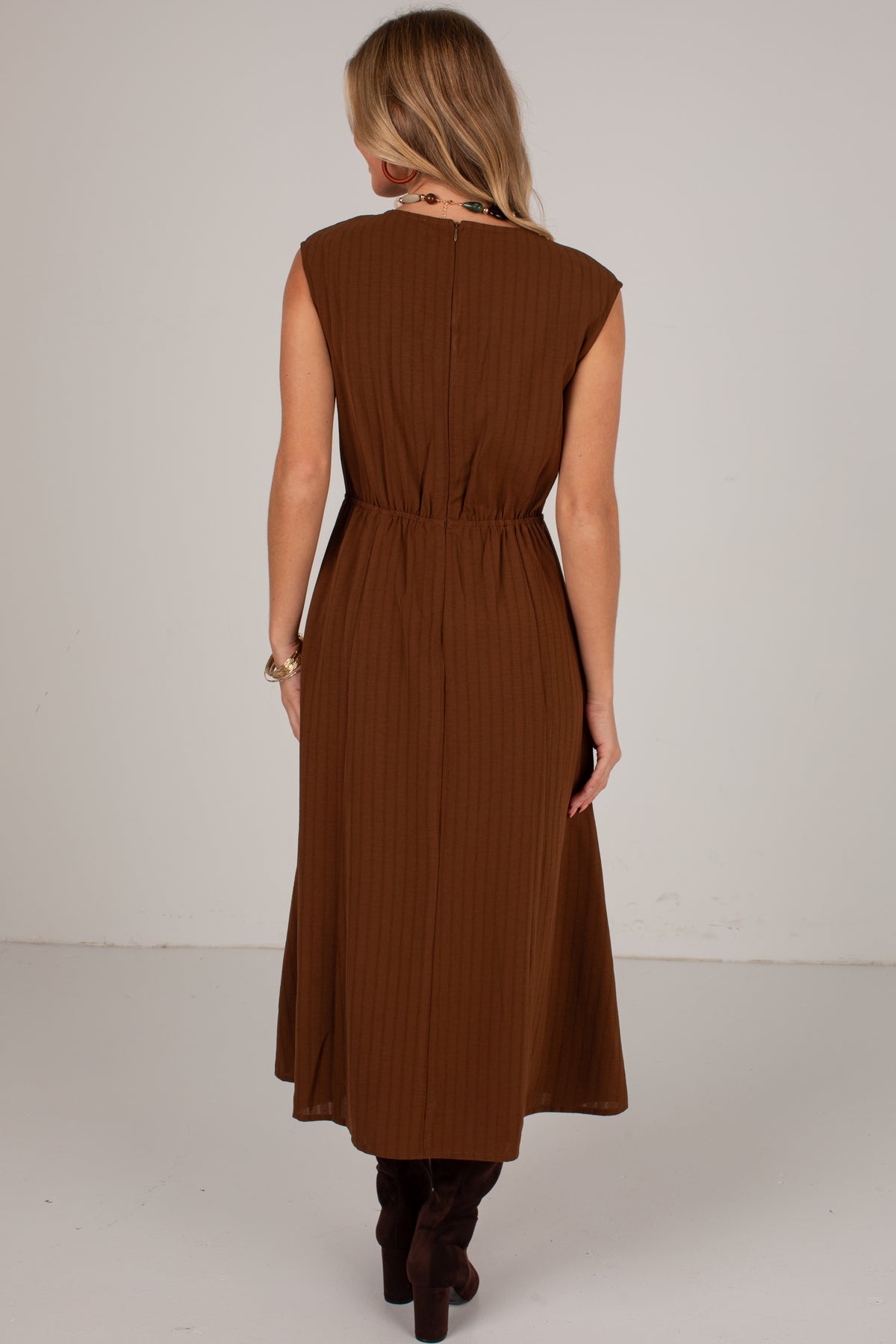 A woman viewed from behind wearing a sleeveless brown midi dress with a gathered waist and pleated texture, paired with dark brown boots, against a plain light gray studio background.