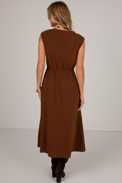 A woman viewed from behind wearing a sleeveless brown midi dress with a gathered waist and pleated texture, paired with dark brown boots, against a plain light gray studio background.