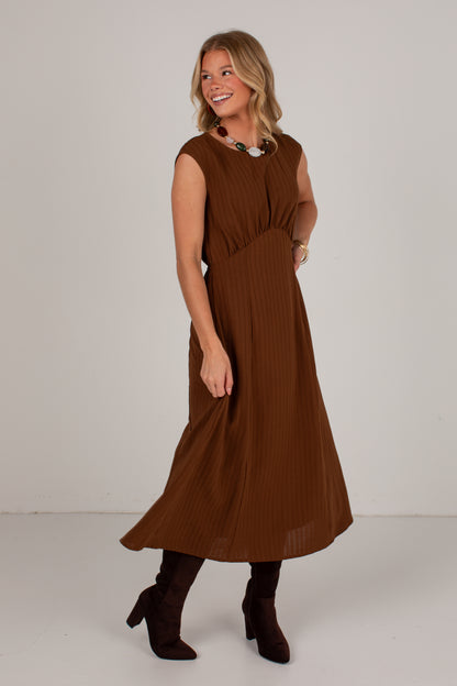 A woman modeling a sleeveless brown midi dress with a gathered waist and pleated texture, paired with dark brown ankle boots, against a plain light gray studio background.