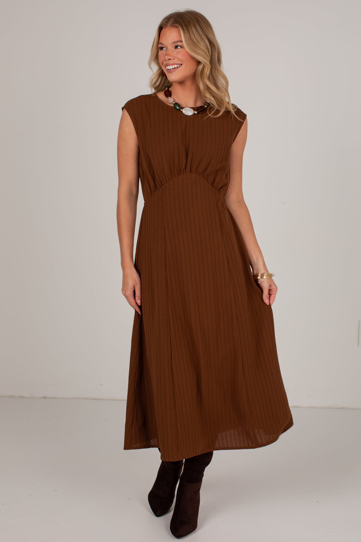 A woman modeling a sleeveless brown pleated midi dress with a gathered waist, paired with dark brown ankle boots and a chunky statement necklace, set against a plain light gray studio background.
