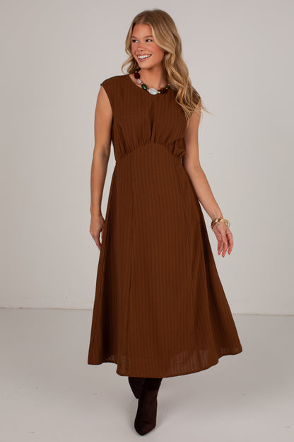 A woman modeling a sleeveless brown midi dress with a gathered waist and pleated skirt, standing against a plain light gray studio background.