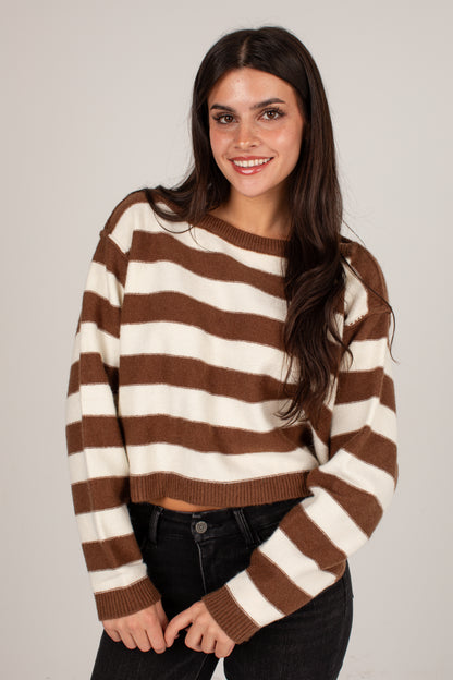 A woman modeling a brown and cream striped cropped knit sweater with black jeans, photographed against a plain light gray studio background.