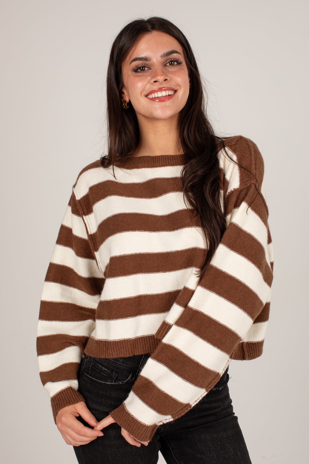 A smiling young woman wearing a brown and cream horizontally striped knit sweater with balloon sleeves, paired with black jeans, against a solid light gray background.