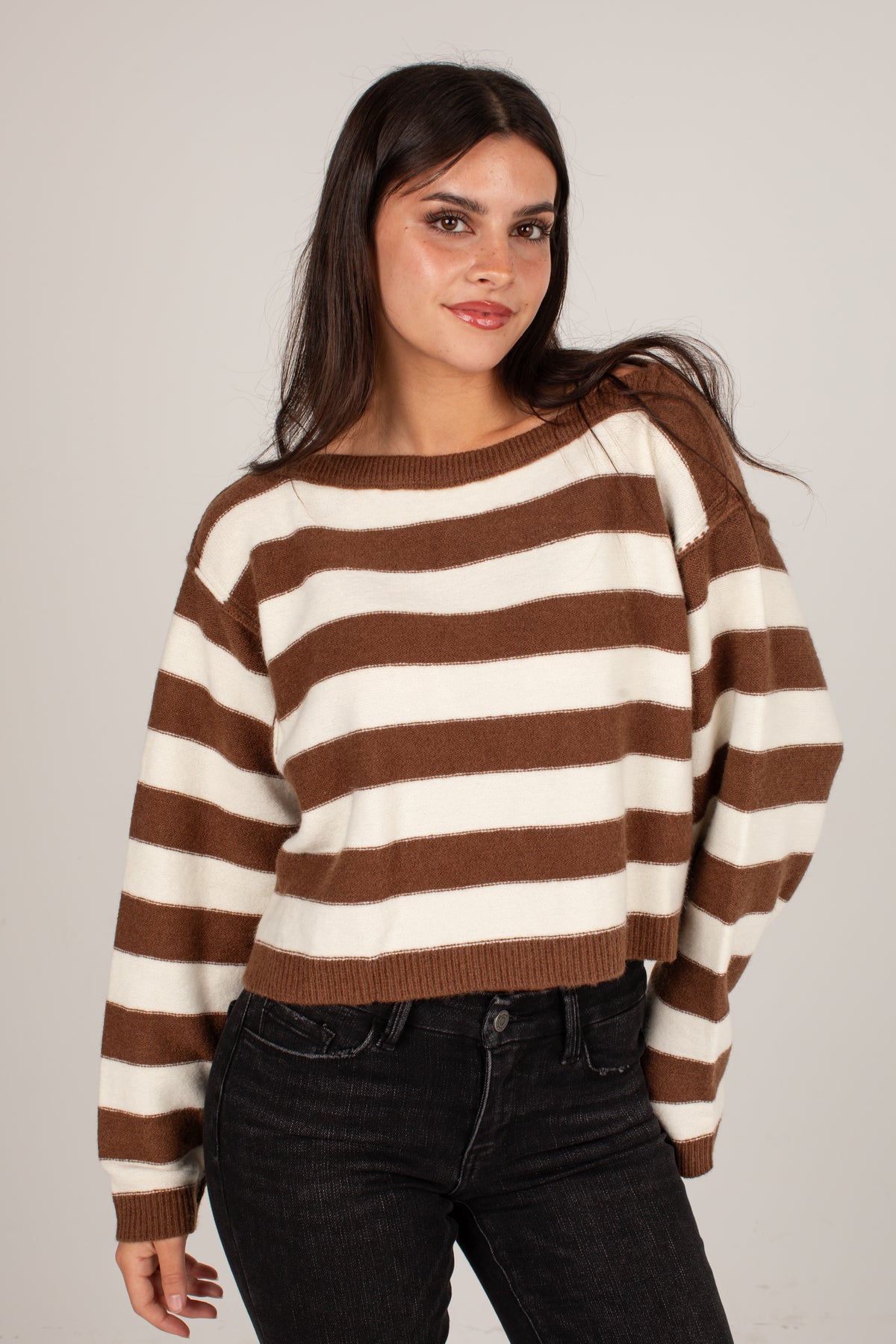 A woman wearing a brown and cream striped knit sweater with black jeans, standing against a plain light gray studio background.