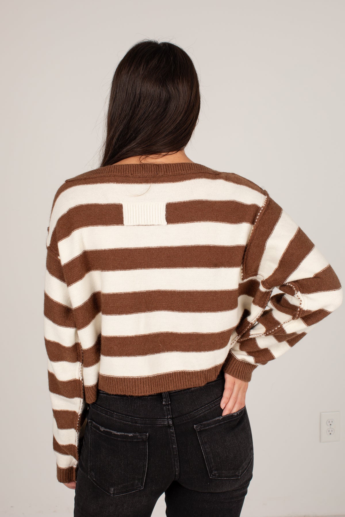 A woman viewed from behind wearing a brown and cream striped knit sweater with balloon sleeves and dark wash jeans, standing against a plain light gray wall.