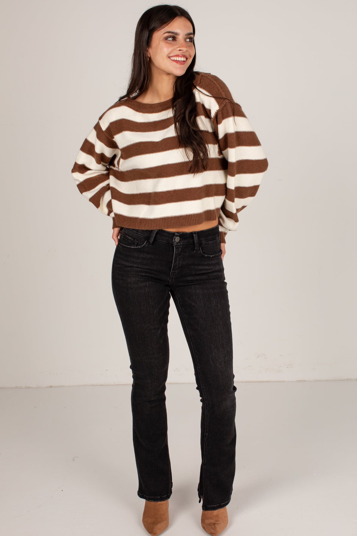 A woman modeling a brown and cream striped cropped knit sweater with balloon sleeves, paired with black jeans and tan boots, against a plain light gray studio background.