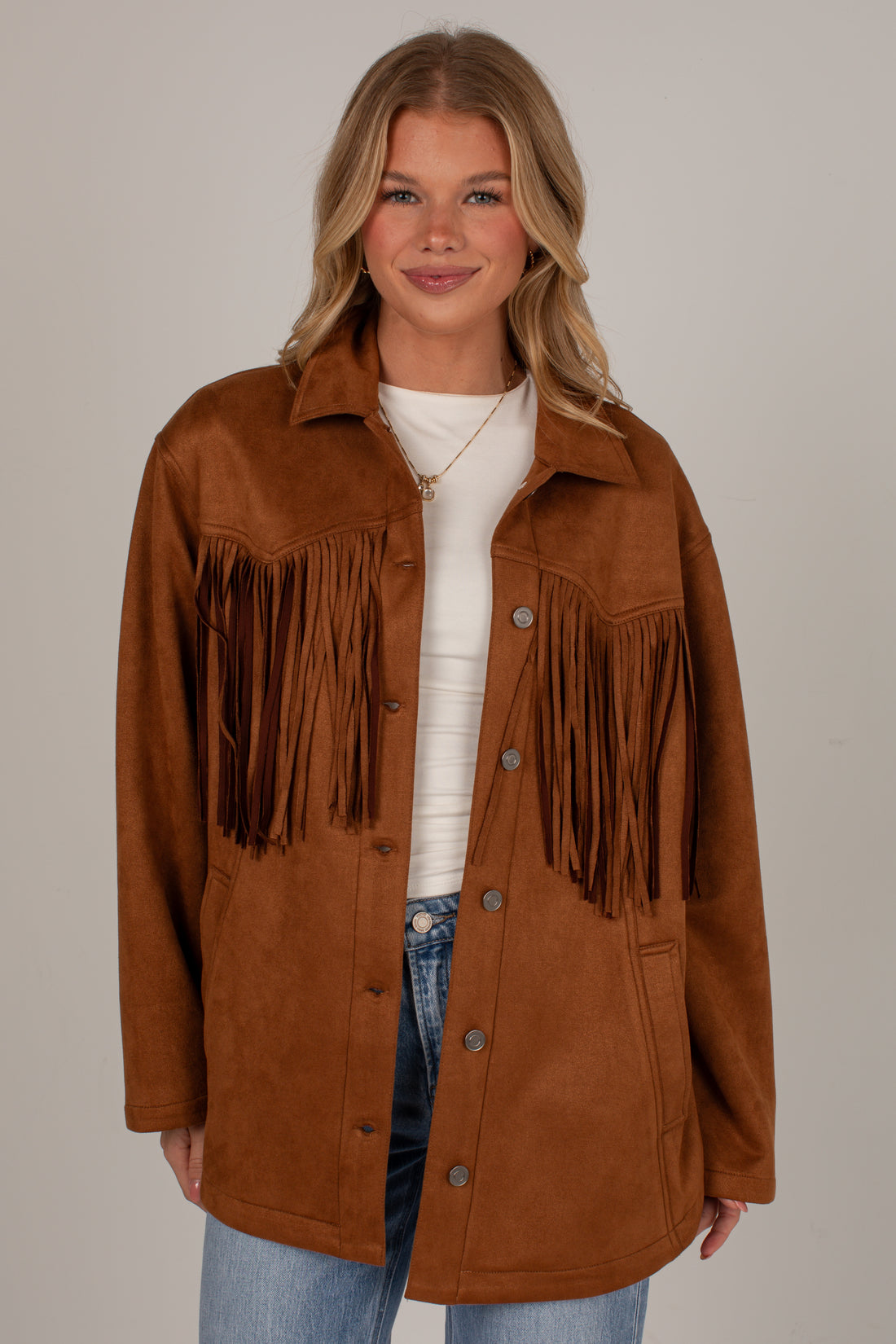 A woman modeling a brown faux-suede fringe jacket over a white top and blue jeans, photographed against a plain light gray studio background.