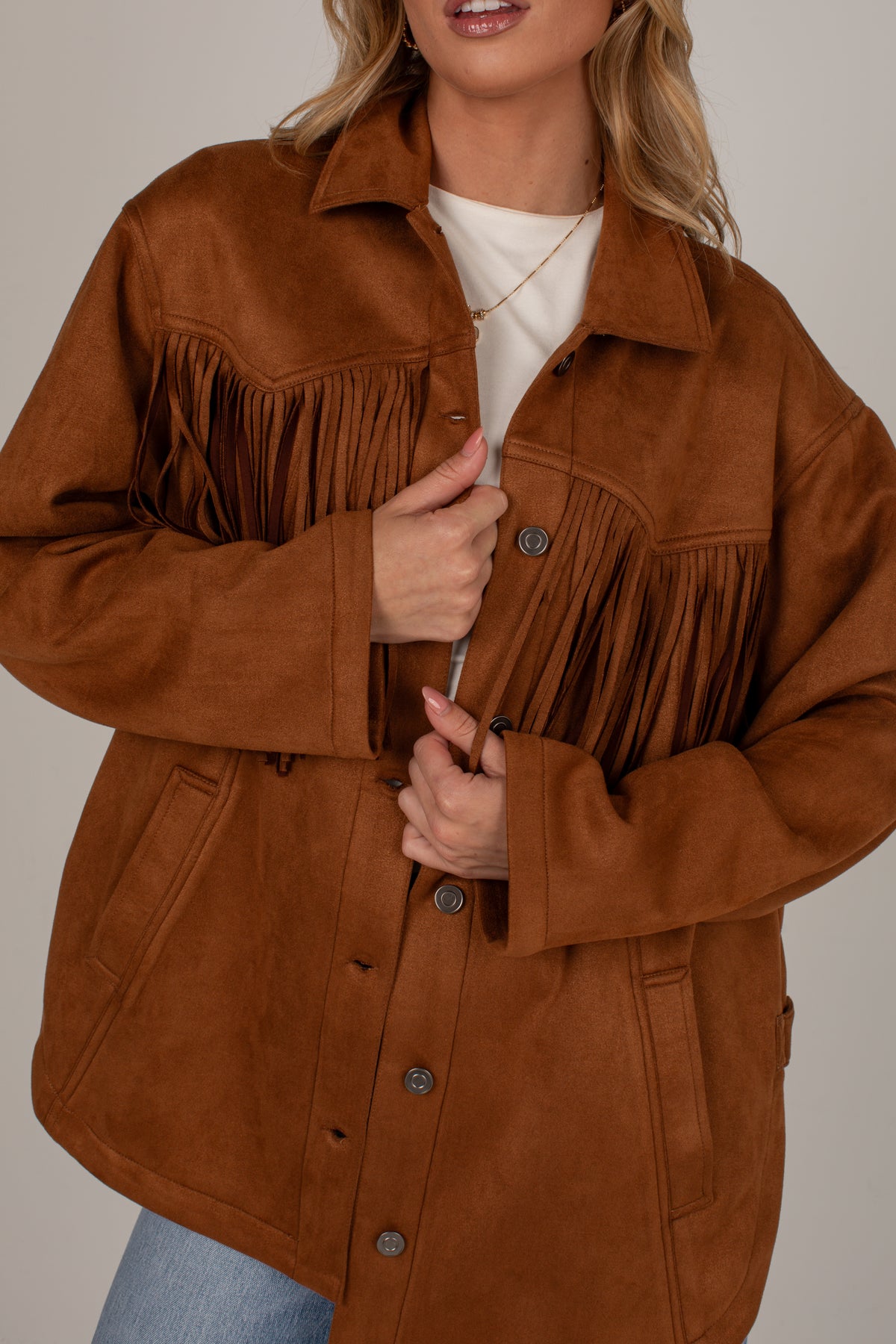 A woman wearing a brown suede-like fringed jacket over a white top and blue jeans, shown from the shoulders to the hips against a plain light gray studio background.