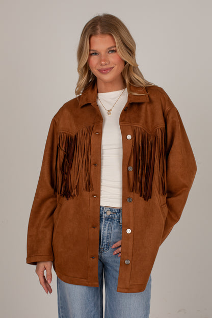 A woman modeling a brown suede fringe jacket over a white ribbed top and blue jeans, photographed against a plain light gray studio background.