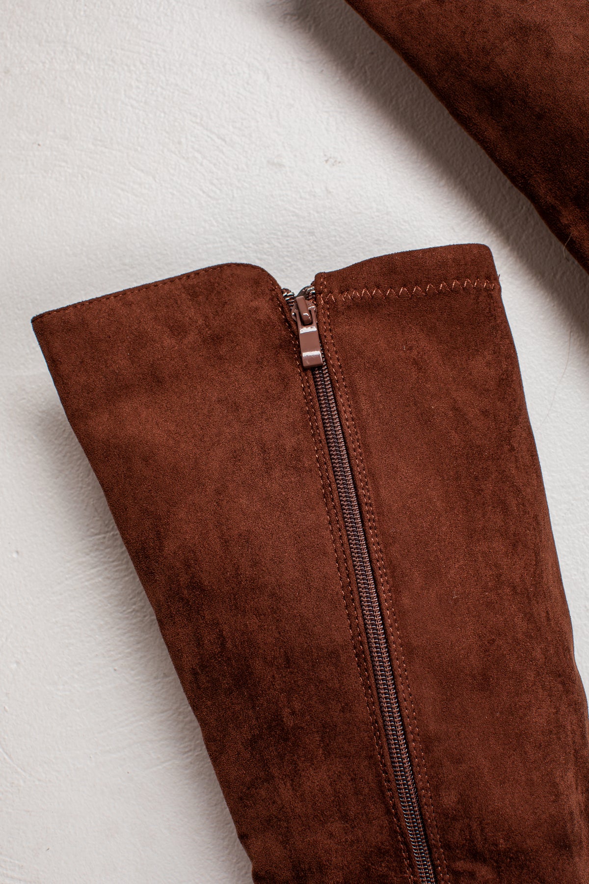 Close-up of a brown suede boot with a dark zipper and a rose gold-toned metal pull tab, laid flat on a textured off-white surface.