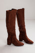 A pair of brown suede knee-high boots with side zippers and low block heels, photographed on a light gray studio background.