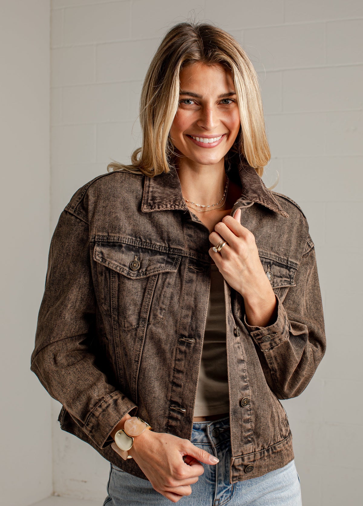 A woman wearing a washed brown denim jacket over a dark top and light blue jeans, standing against a white paneled wall.
