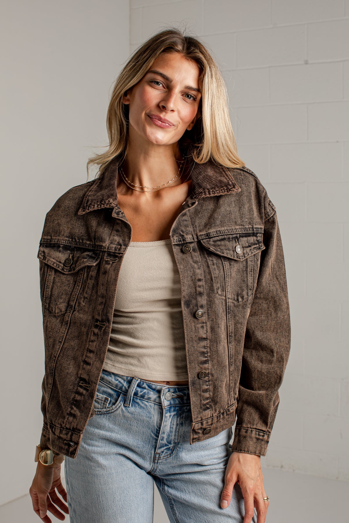 A woman wearing a washed brown denim jacket over a beige tank top and light blue jeans, standing against a light gray brick wall.