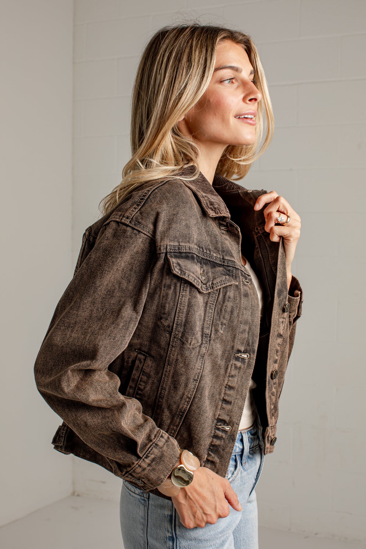 A woman wearing a washed brown denim jacket over a white top and light blue jeans, standing in a bright white studio setting.
