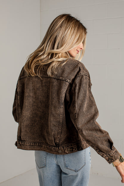 A woman wearing a washed dark brown cropped denim jacket and light blue jeans, shown from the back and side against a white brick wall.