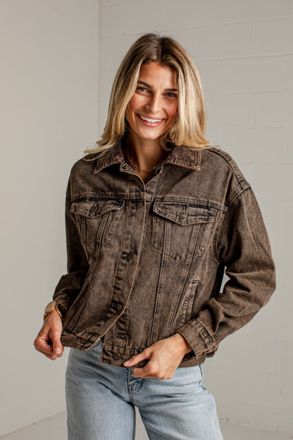 A woman wearing a dark brown acid-wash denim jacket and light blue jeans, standing against a white brick wall.