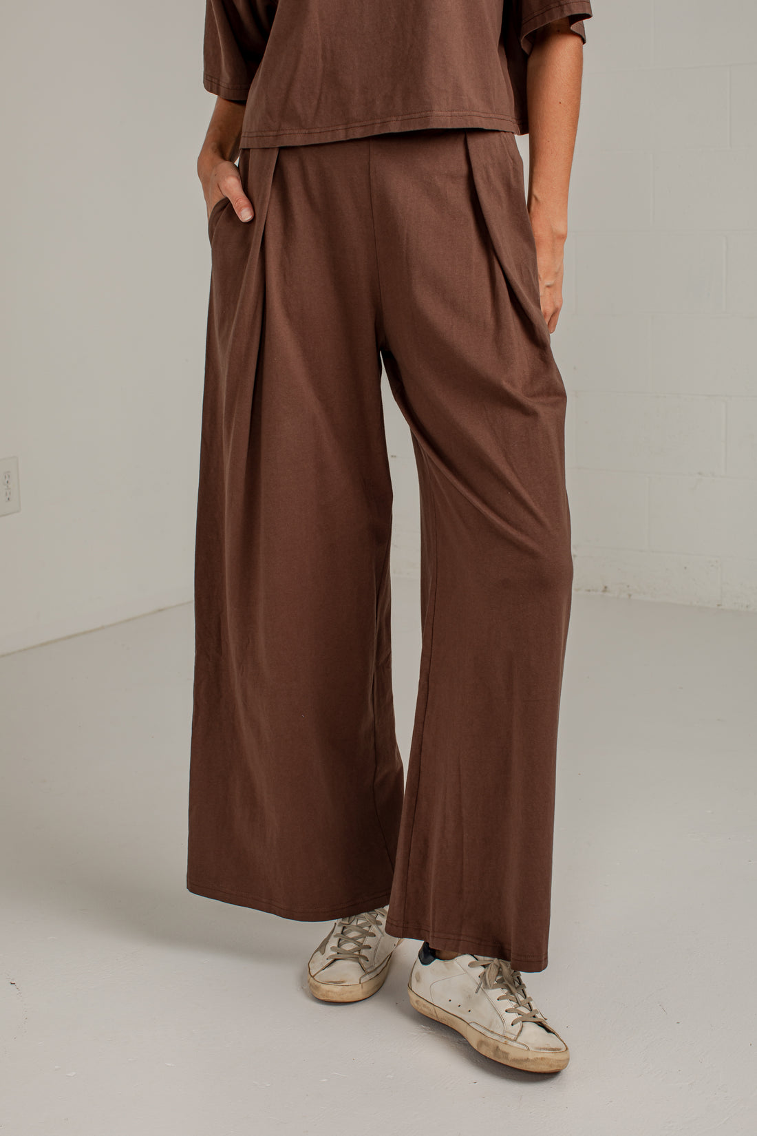 A person wearing loose, wide-leg brown pants with a matching brown top and light-colored sneakers, standing in a plain indoor setting with a white wall and floor.