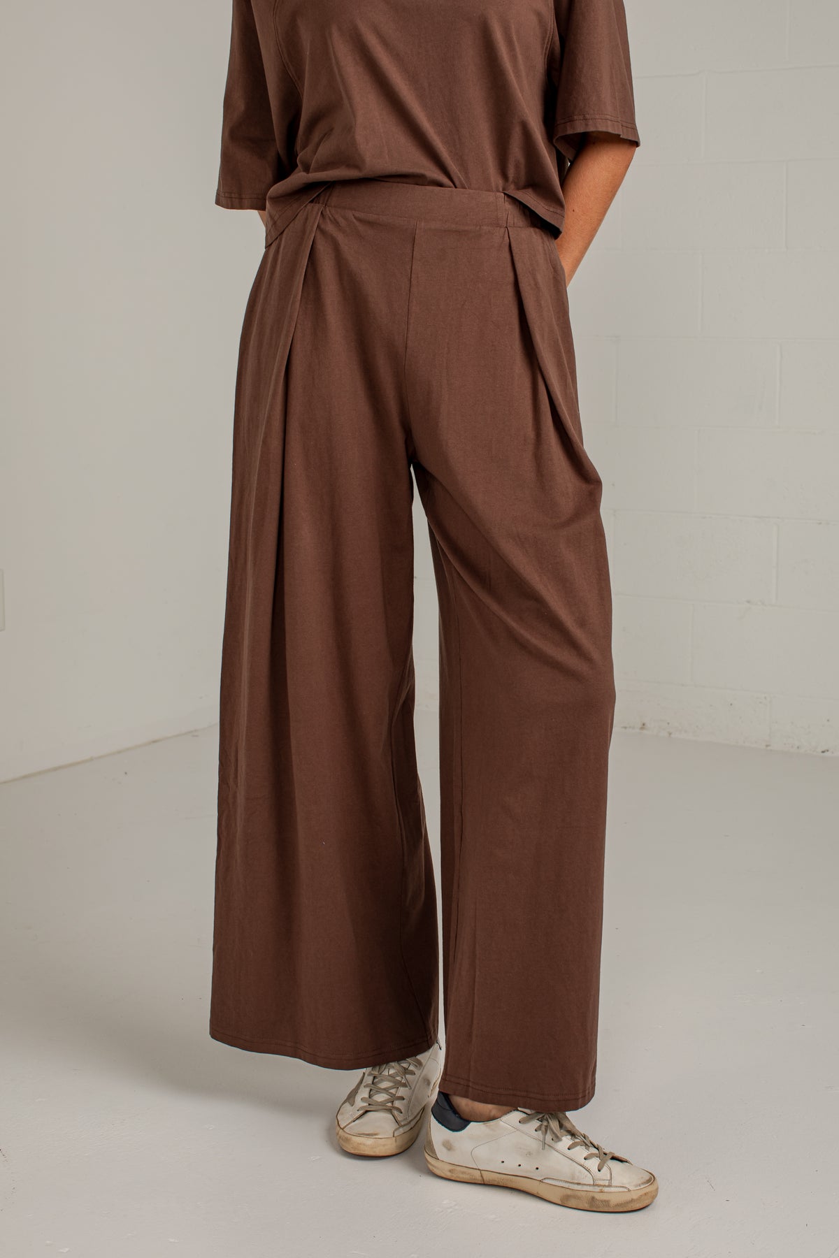 A person wearing brown wide-leg pleated trousers with a matching brown top and white sneakers, shown from the shoulders down in a studio setting.