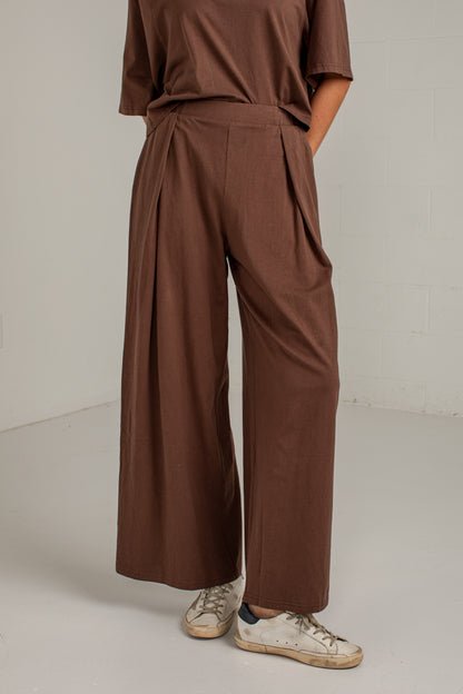 A person wearing brown wide-leg pleated trousers with a matching brown top and white sneakers, shown from the shoulders down in a studio setting.