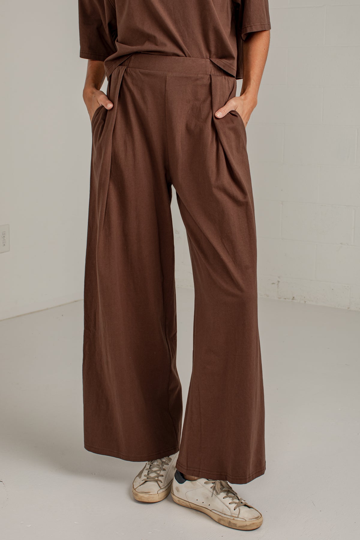 A person wearing brown wide-leg pleated pants with a matching brown top and distressed white sneakers, standing in a minimalist studio setting.