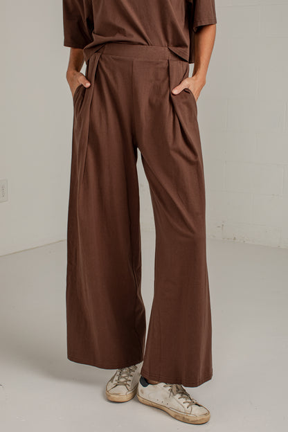 A person wearing brown wide-leg pleated pants with a matching brown top and distressed white sneakers, standing in a minimalist studio setting.