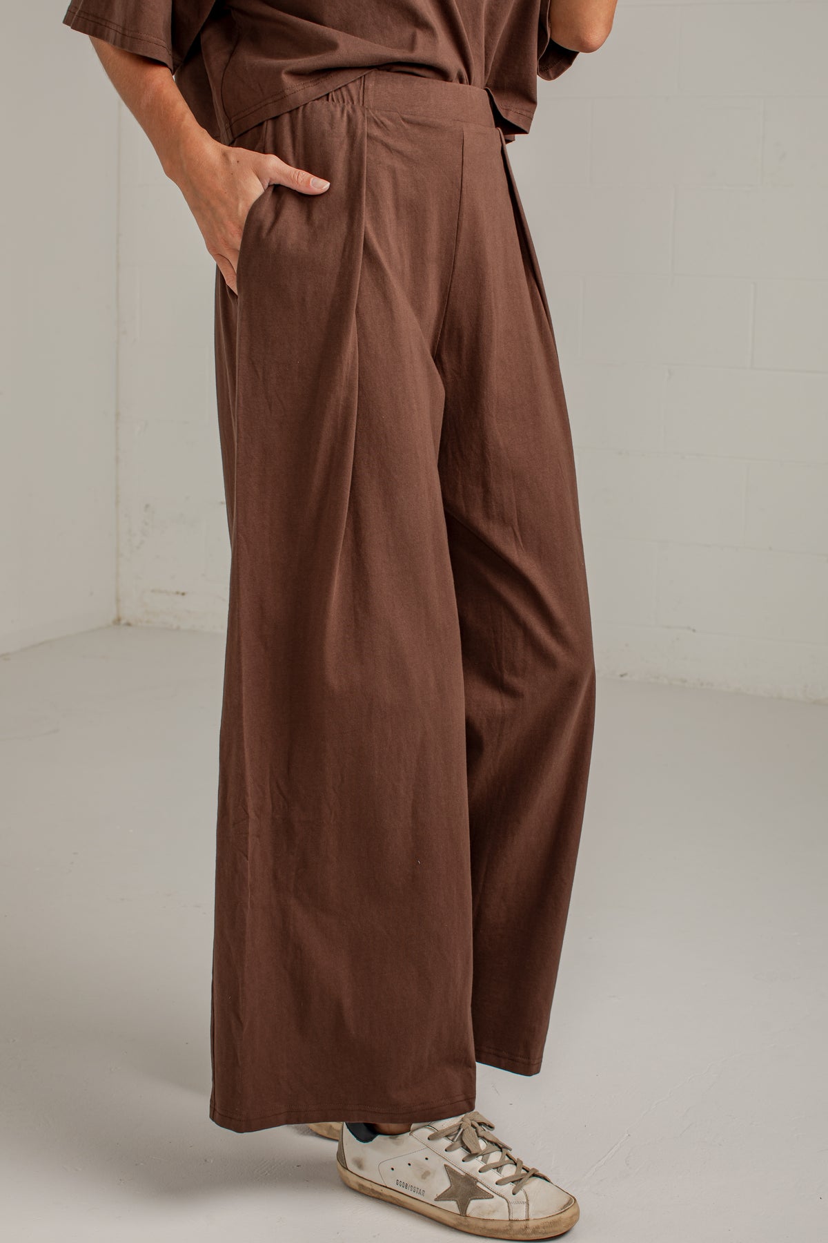 A person wearing loose, wide-leg brown pants with a matching brown top and white sneakers, standing in a plain indoor setting with a white wall and floor.
