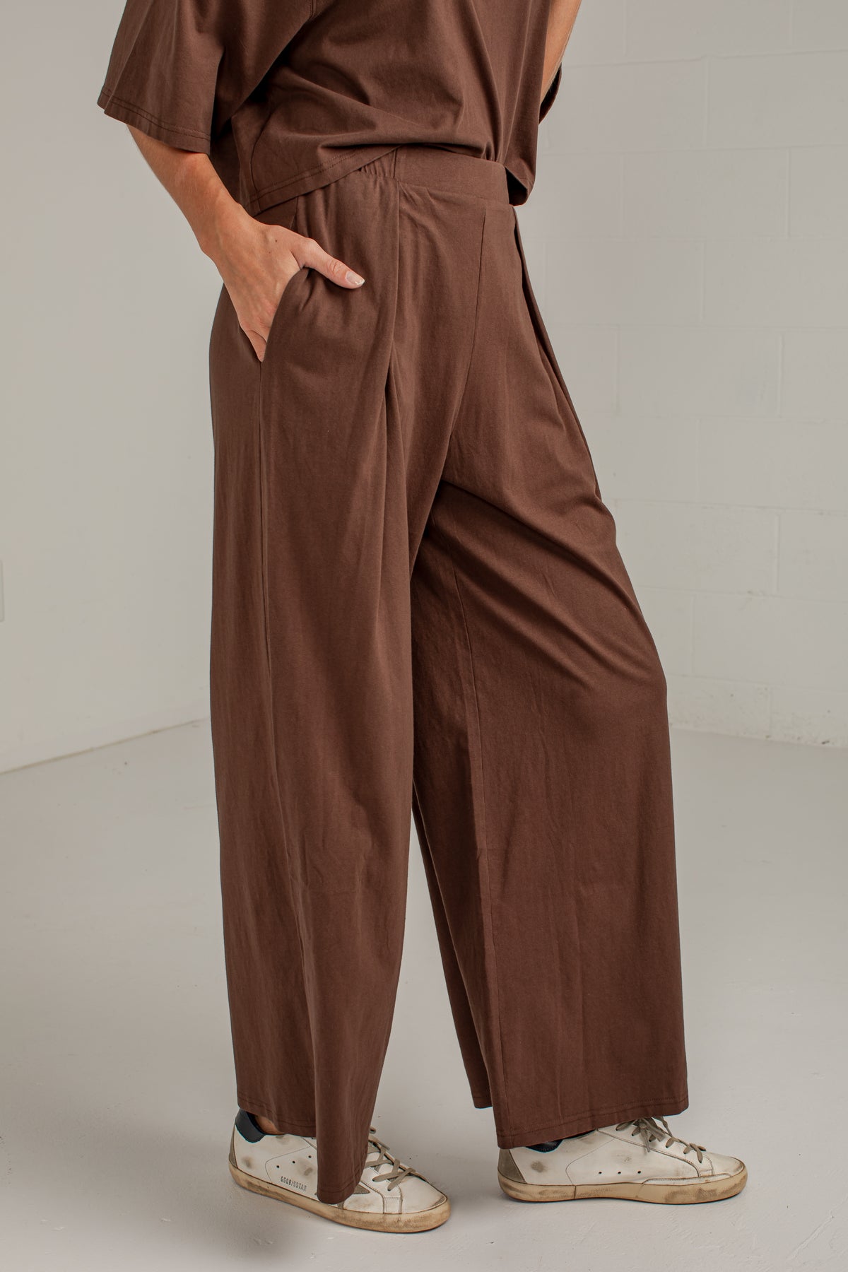 A person wearing loose, wide-leg brown pants with pleats and side pockets, paired with a matching brown top and white sneakers, standing against a plain light gray studio wall.