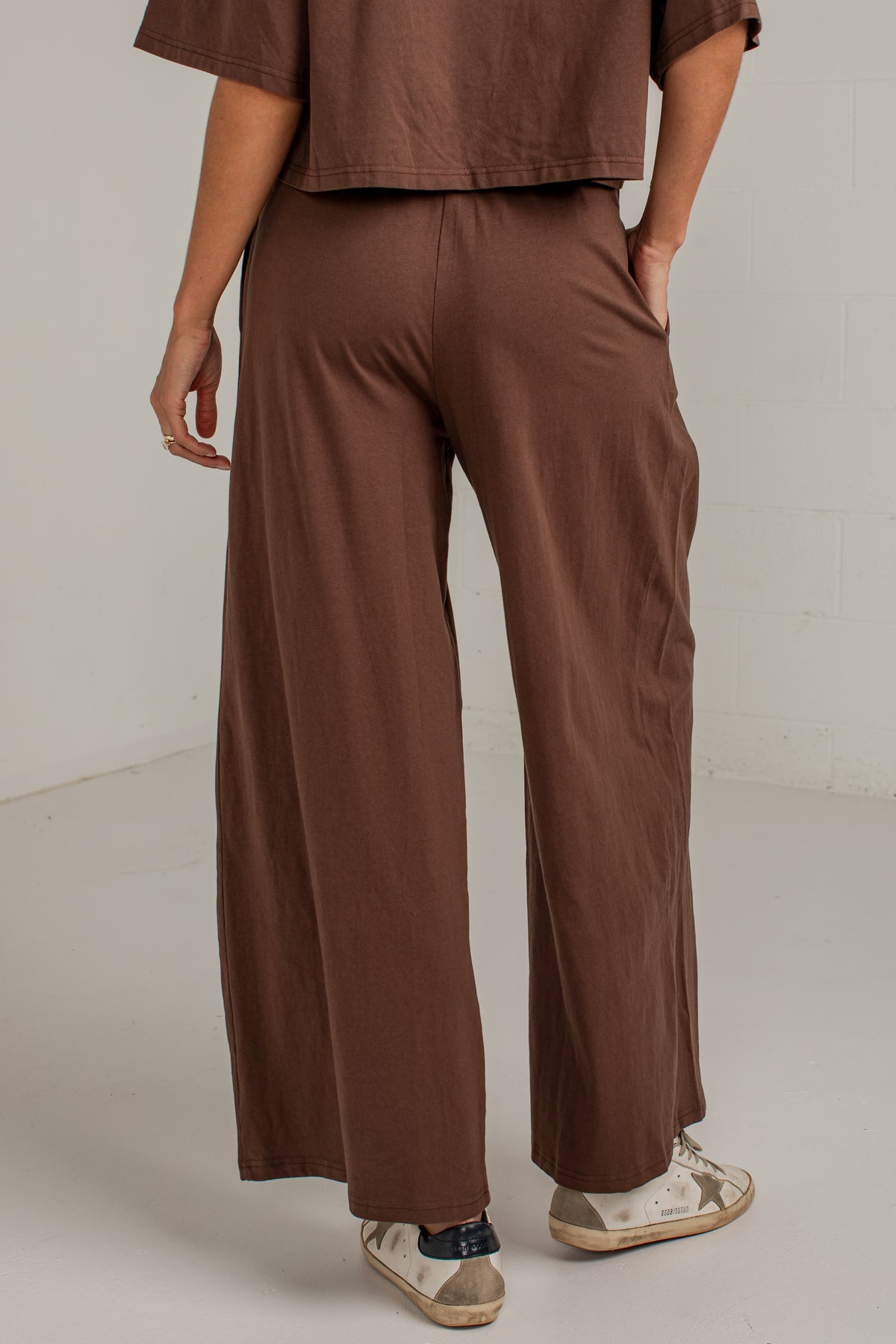 A person shown from the back wearing loose, wide-leg brown pants and a matching brown cropped top, standing in a plain indoor studio setting with sneakers.